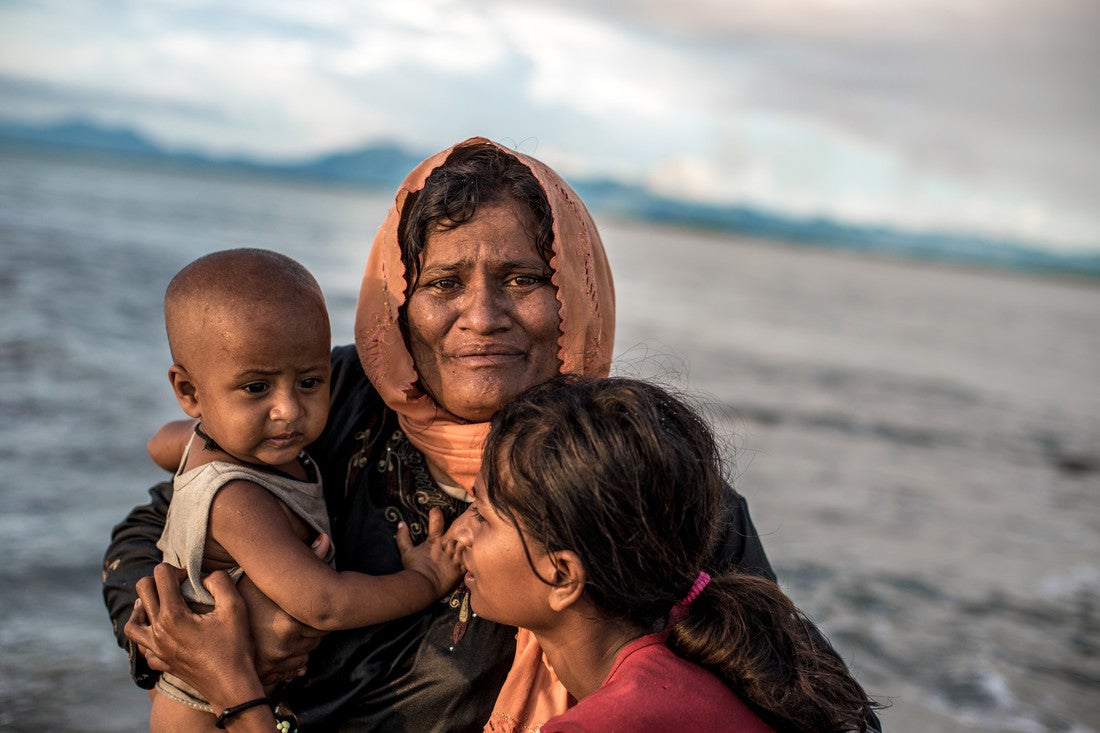 Uma mulher angustiada se amontoa com dois filhos, um dos quais é um bebê, em frente ao oceano.