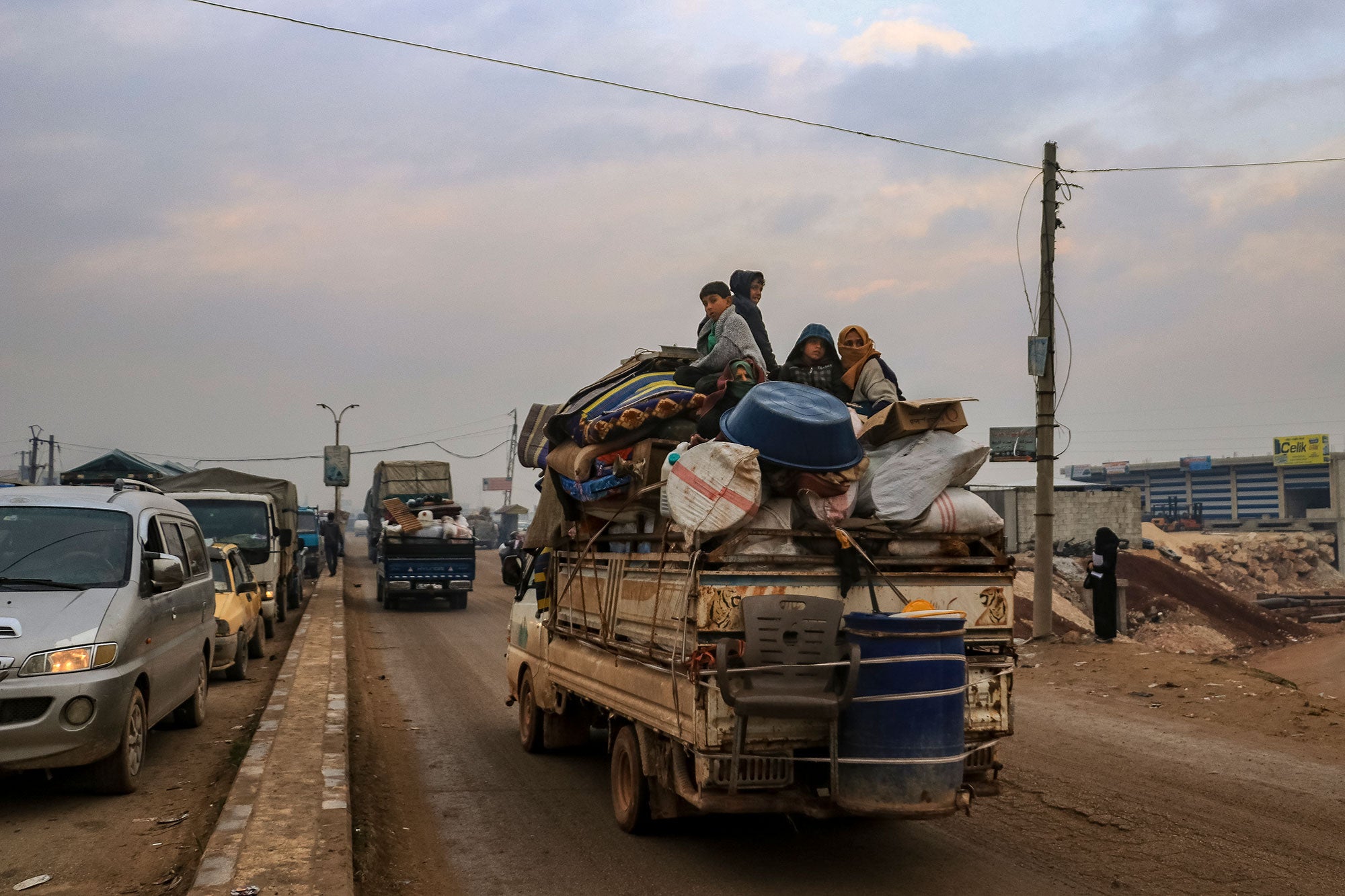Four people sit on top of a truck overflowing with luggage. A chair and large bucket are tied to the back of the truck, which is driving behind a similarly packed van on a dirt road.