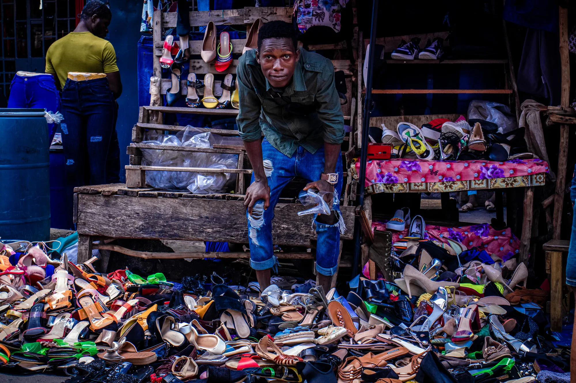 A man stands in front of bundles of shoes in an open-air stall.