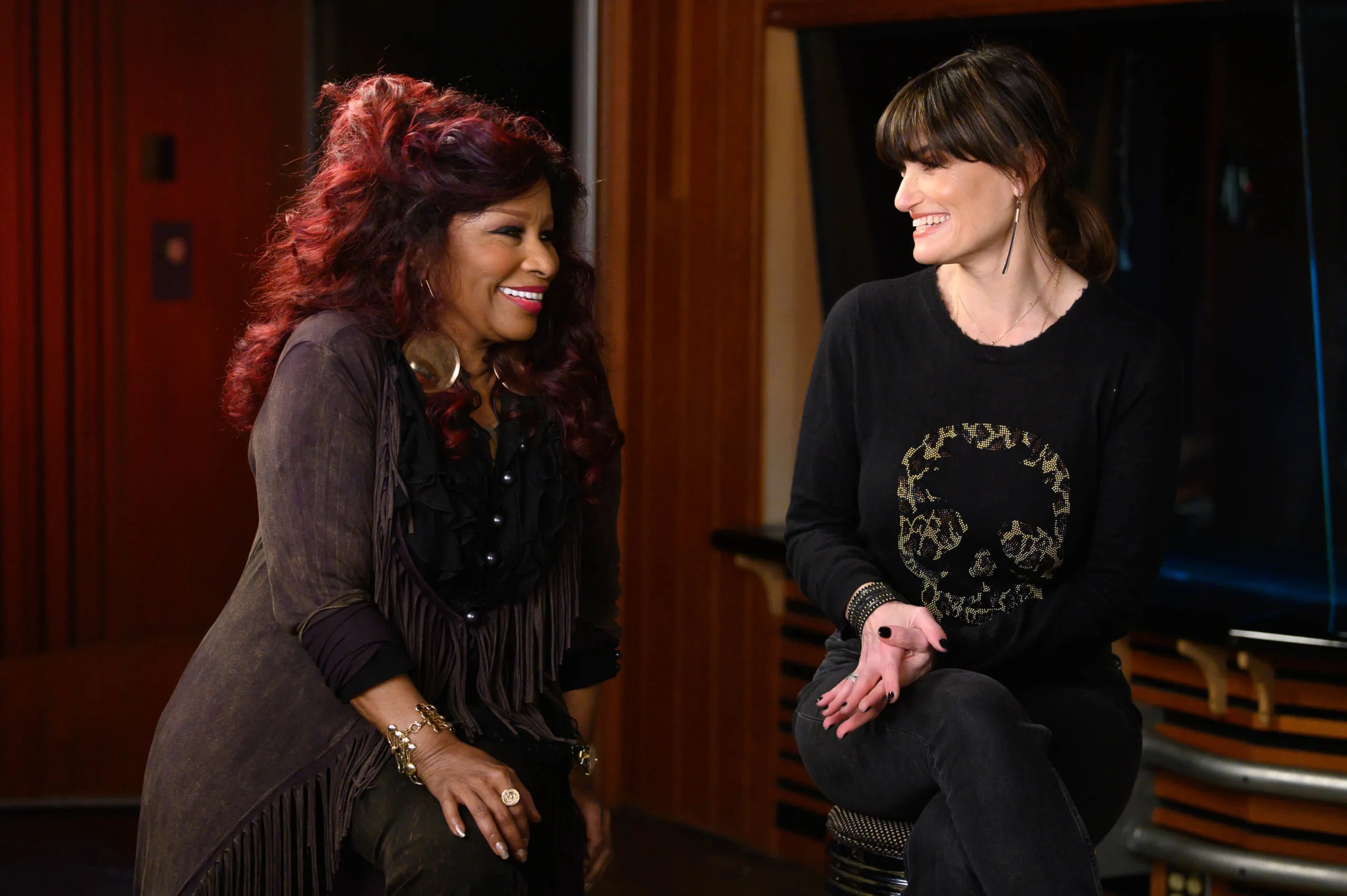 Chaka Khan and Idina Menzel smile widely as they sit next to each other on stools in a recording booth.