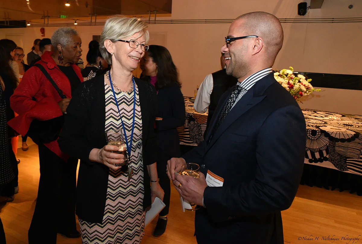 Ambassador Kristi Kauppi is talking to a man in a black suit. Both are holding glasses of a beverage