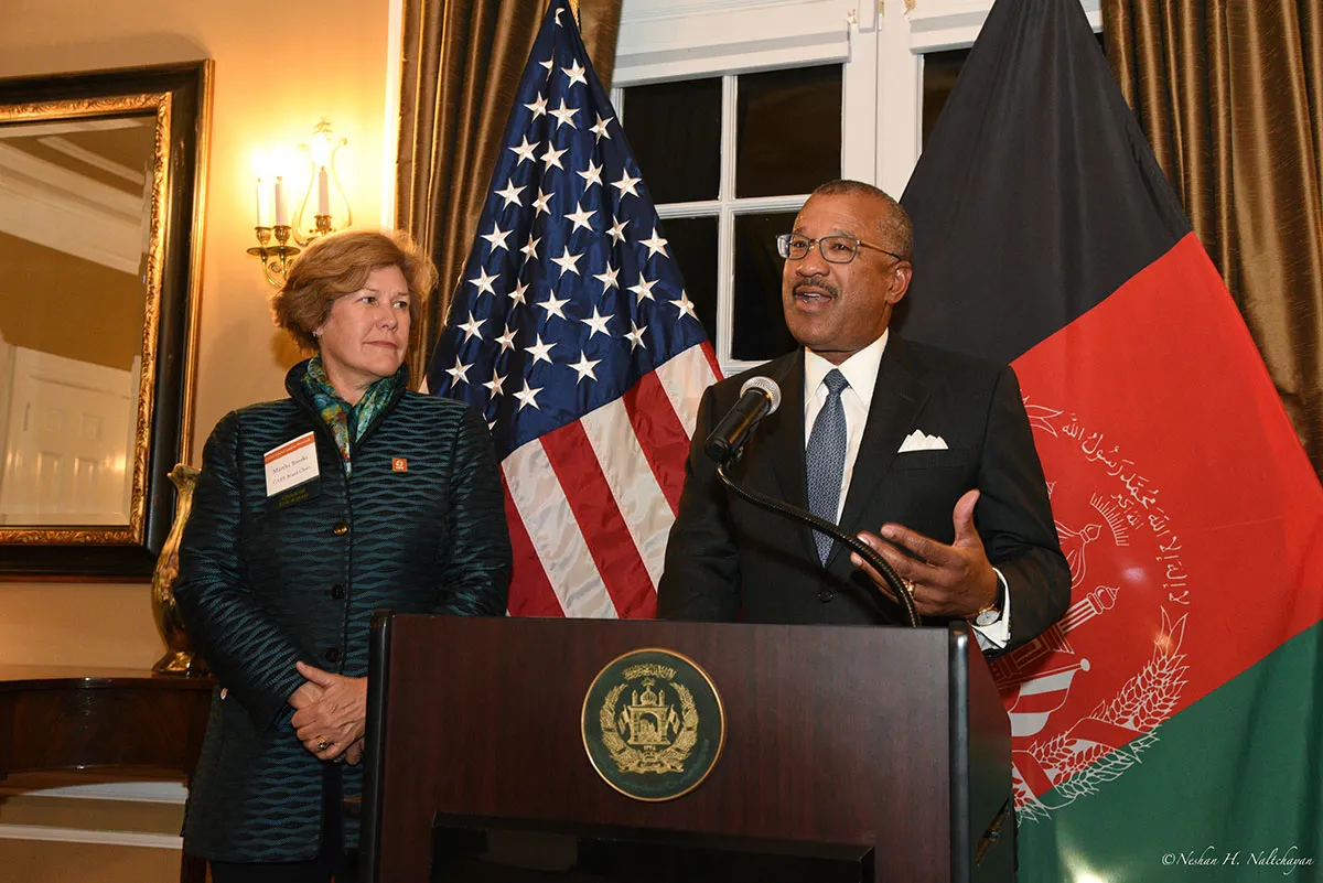 A man in a black suit is giving a speech next to a woman in a dark green suit with the US and Afghan flags in the background