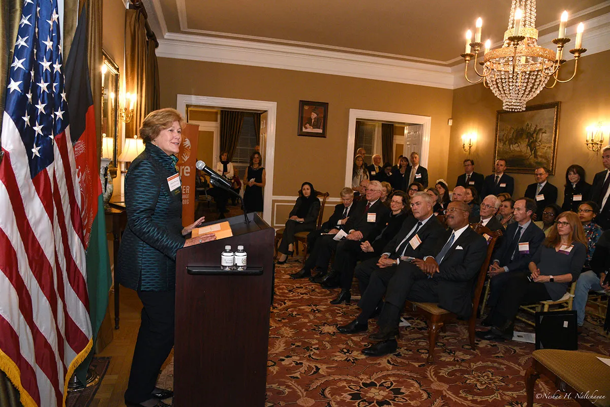 A woman in a dark green suit is holding a speech in front all guests who are sitting on chairs