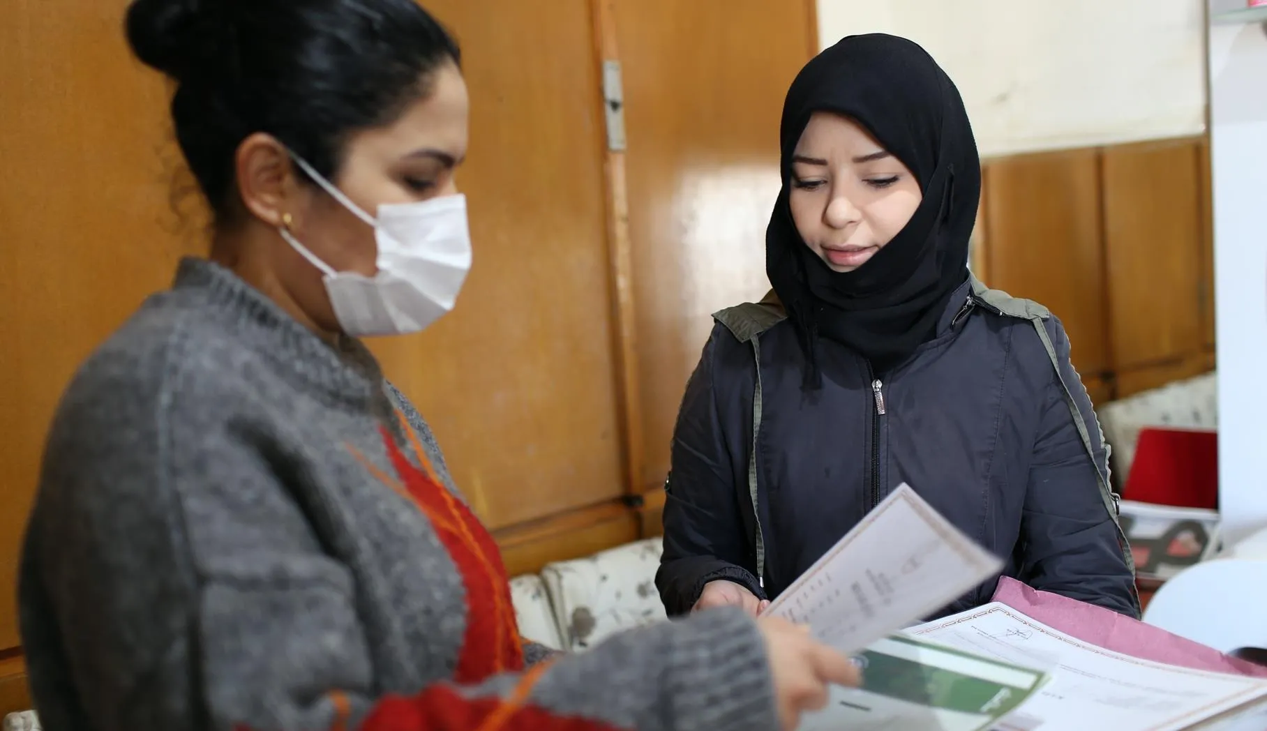 A woman wearing a black hijab approaches another woman, who is wearing a facemask and looking through papers.