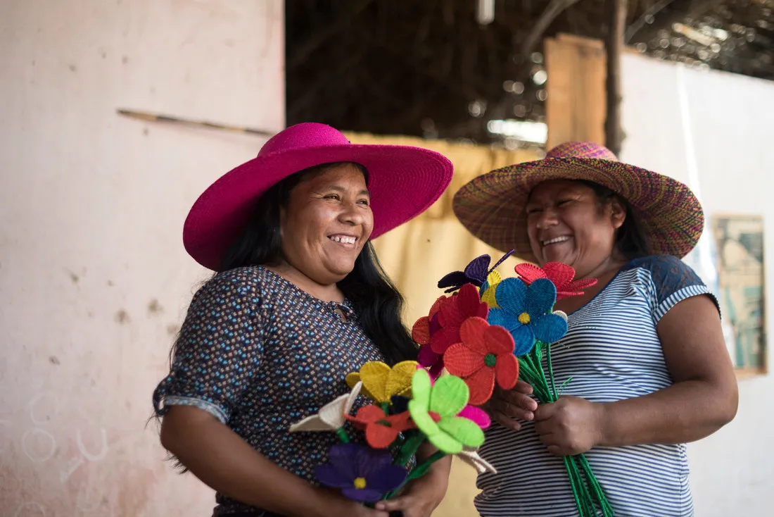 Two women wearing large wide-brimmed hats smile while standing next to each other. They're each holding large, brightly colored felt flowers.