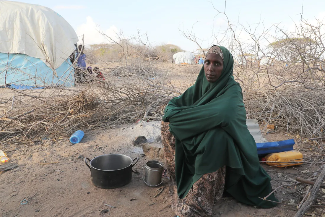 A woman wearing a dark green covering sits on the sandy ground near a tent.