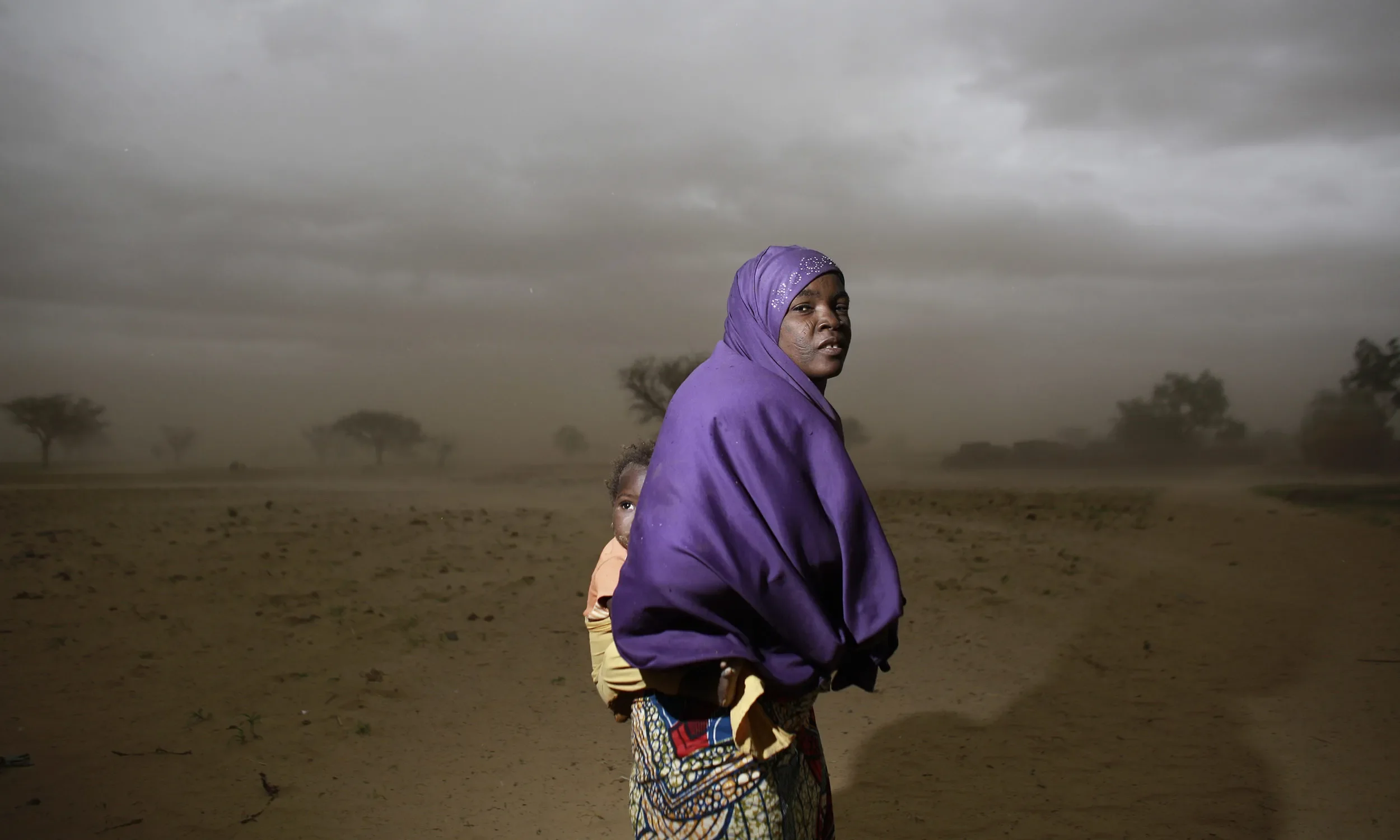 Woman in purple head covering and shawl, carrying a child on her back, looks back toward the camera with a grey landscape in the background
