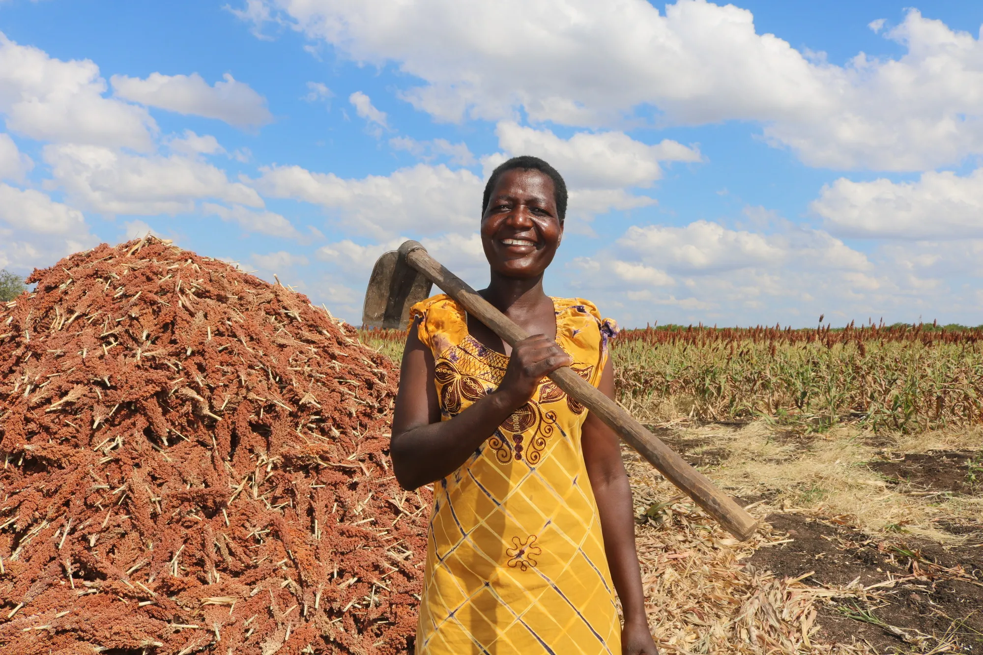 Smiling woman outdoors, holding a tool in front of a stack of harvested crops