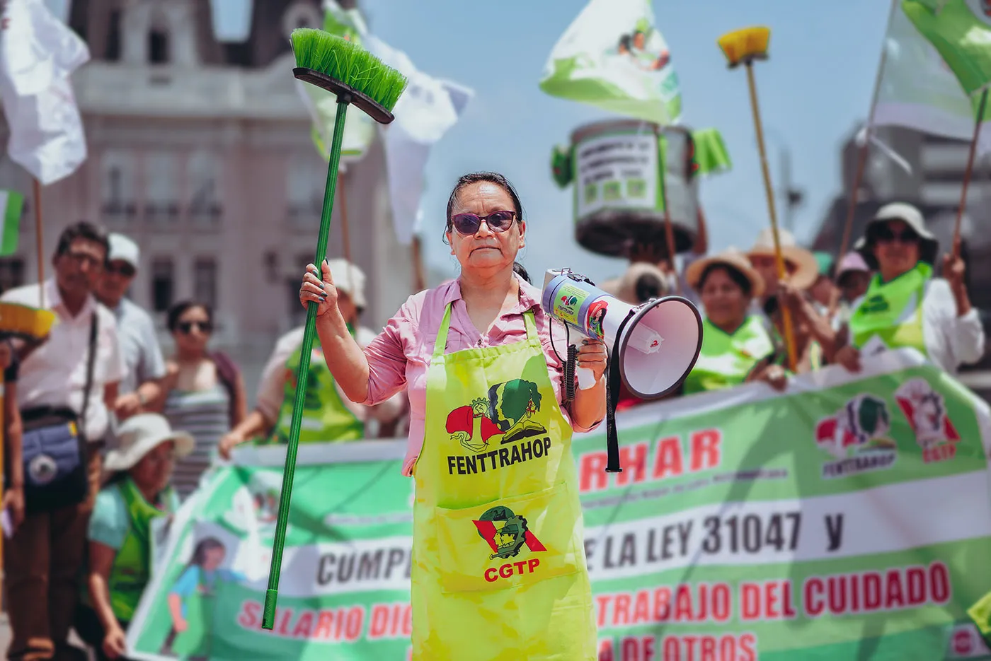 A woman wearing a neon yellow apron leads a march. She's holding a broom in one hand and a megaphone in the other.