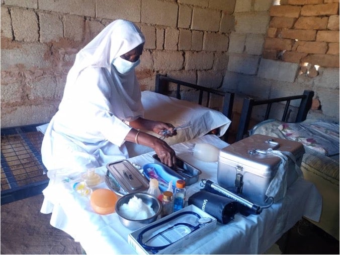 A woman in white, wearing mask and with head covered, next to a table of medical instruments and supplies.