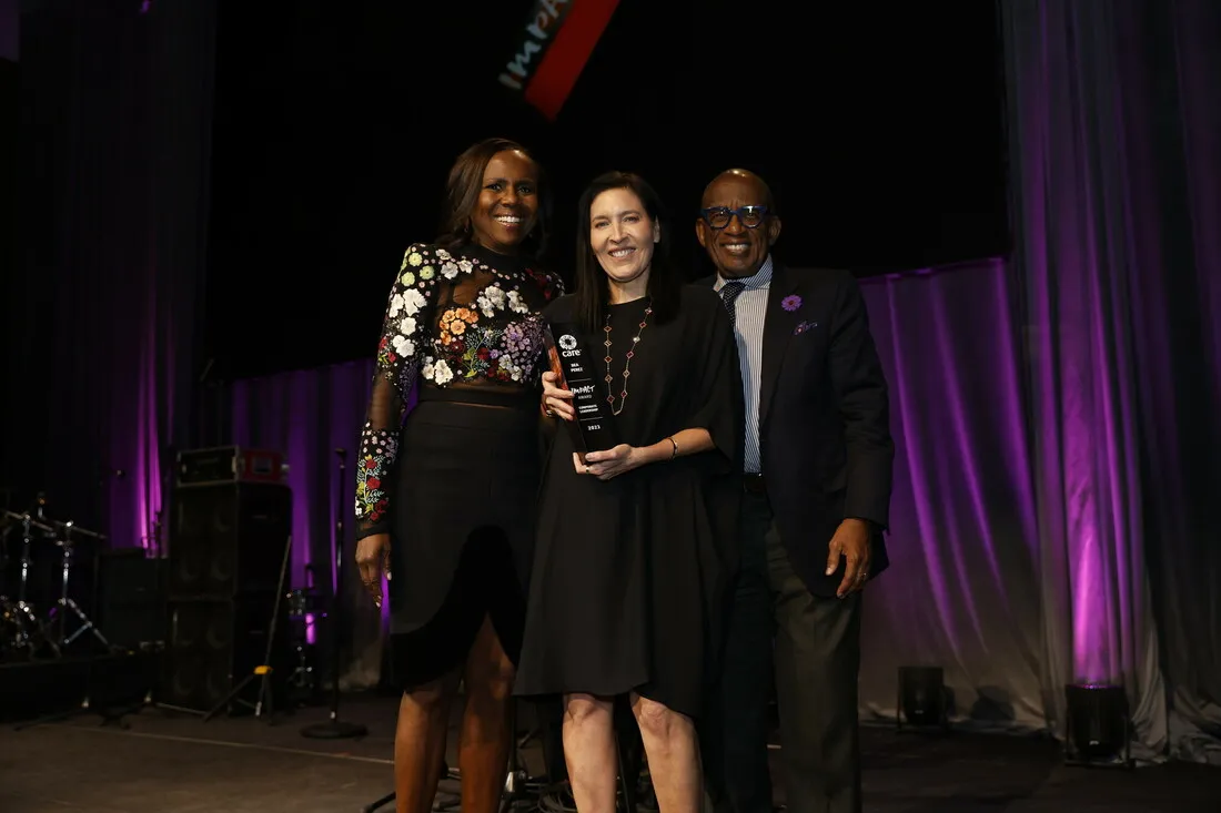 A woman holds up an award while standing in between two hosts.