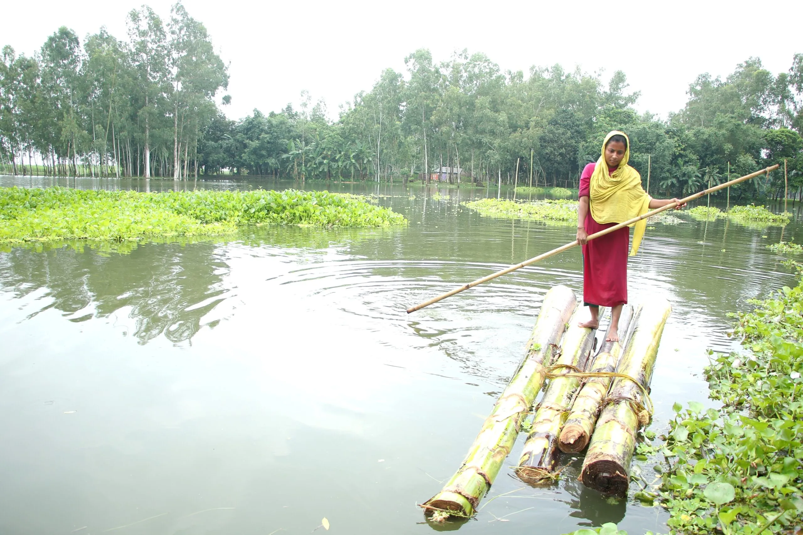 The photograph shows a young woman navigating through a flood-affected area in Bangladesh using a raft made from banana tree trunks.
