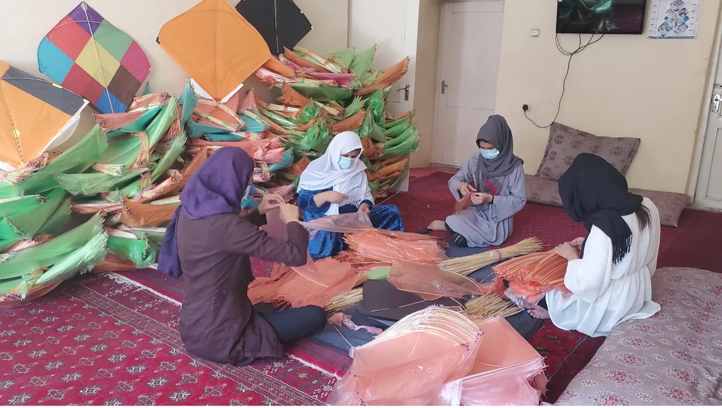 Four women wearing head coverings and masks sit on the floor and work with their hands.
