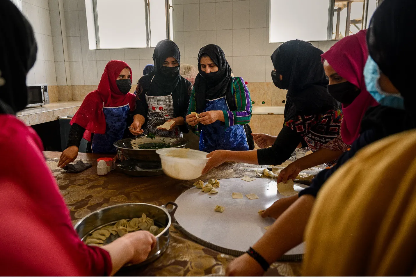 A group of women in head coverings and masks gather around a table, preparing food in bowls and on plates.
