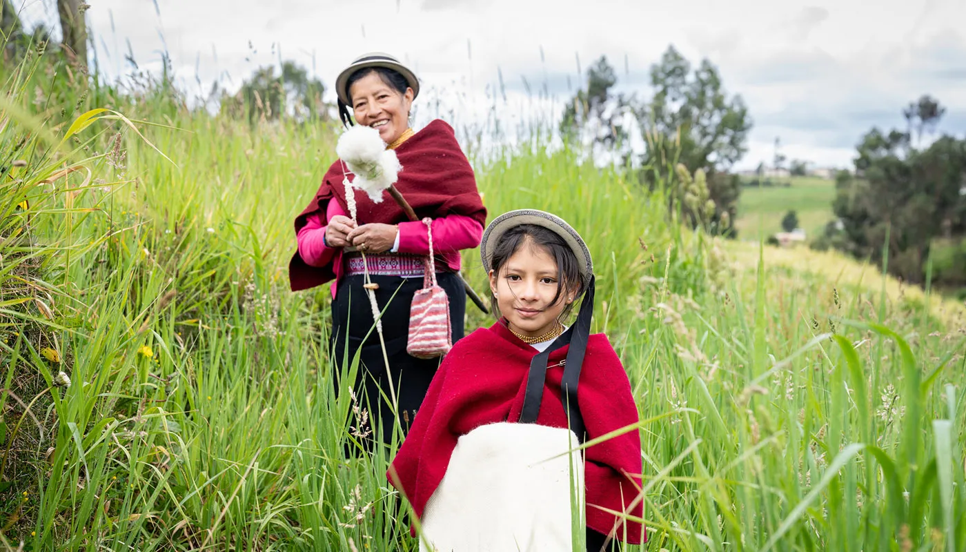 A Honduran woman and girl wearing hats and bright red coats walk through tall grass on a hill.