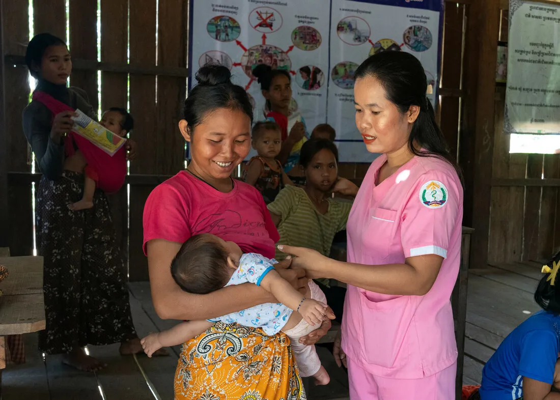 A woman smiles while holding a baby. Another woman, wearing pink scrubs, stands next to her.