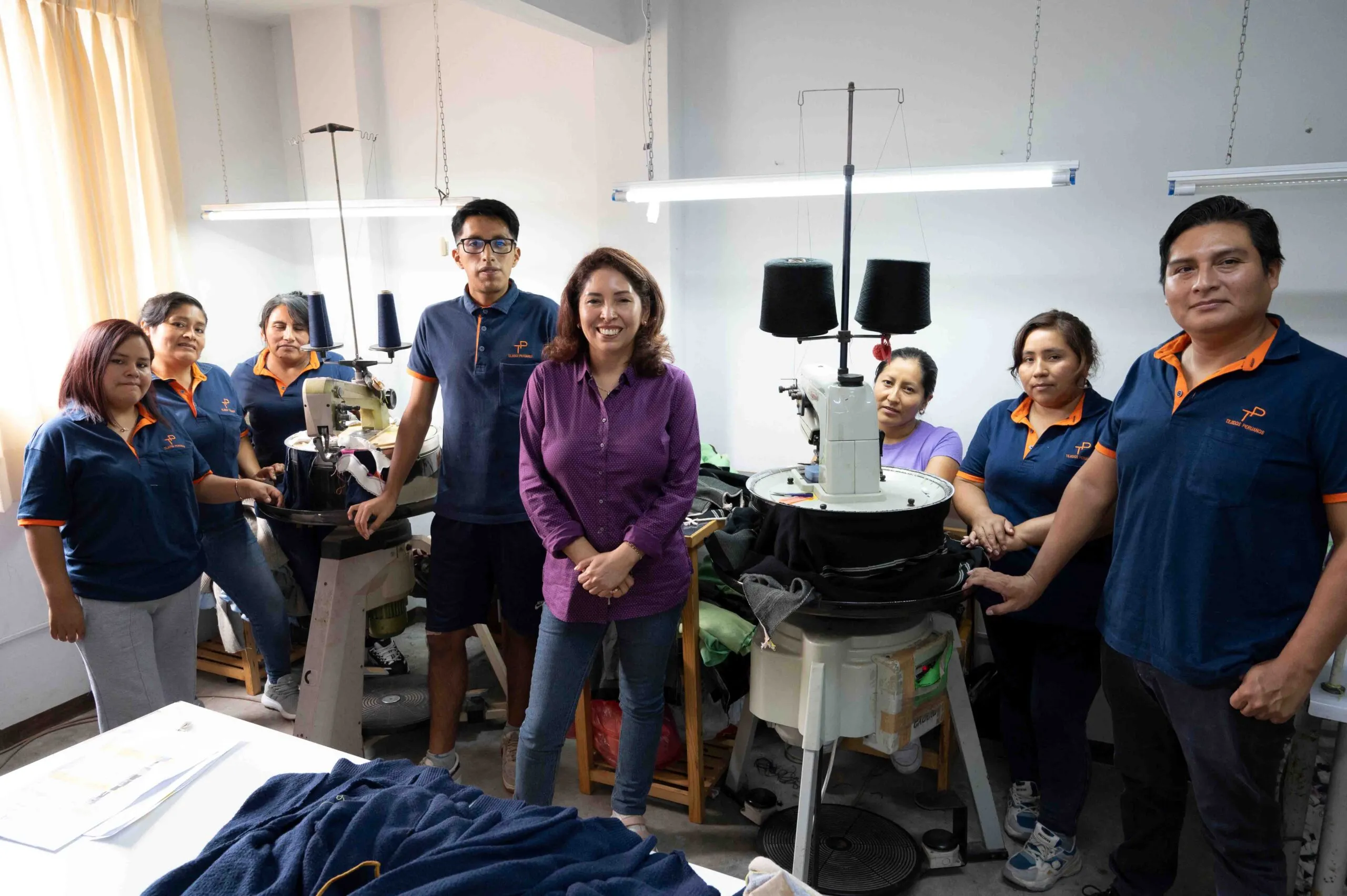 Image of a group of eight people gathered in a room with two sewing machines, a woman in purple at the center.
