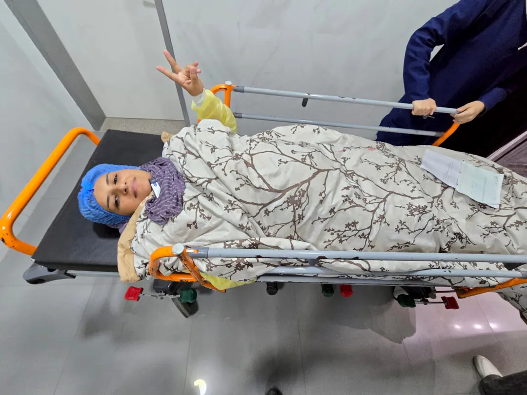 A woman wearing surgical town and head covering, lying on a gurney and holding up two fingers on her left hand.