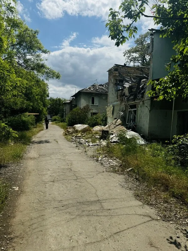 Image of a sunny street, with partially destroyed homes on the right side and a person walking in the distance.