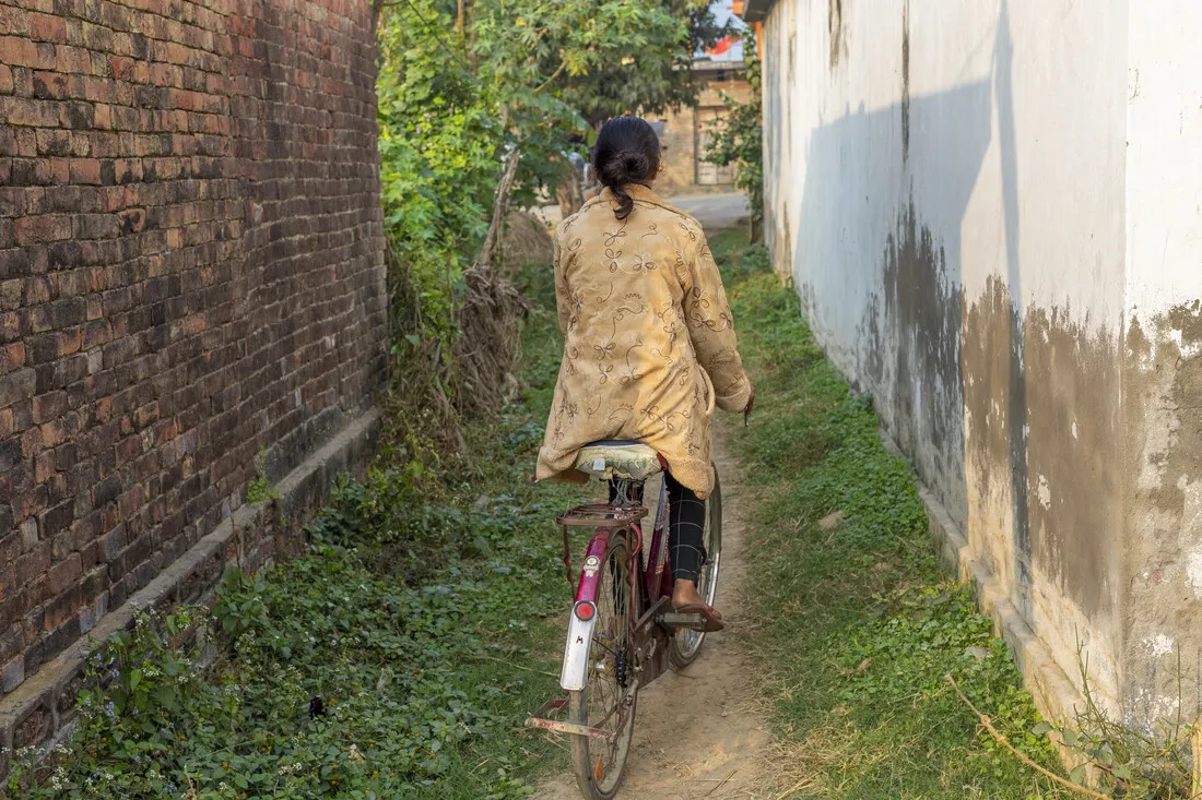 A Nepalese girl, captured from behind by a photographer, rides a bicycle along a narrow alleyway between two houses. She wears a traditional Nepalese salwar kameez. The path is lined with grass on both sides, and the walls are distinctive: one is whitewashed, while the other retains its natural brick color. The girl rides alone, likely heading towards a village road to reach her destination. The background features the road and another house on the opposite side, providing a sense of depth and context to the scene.