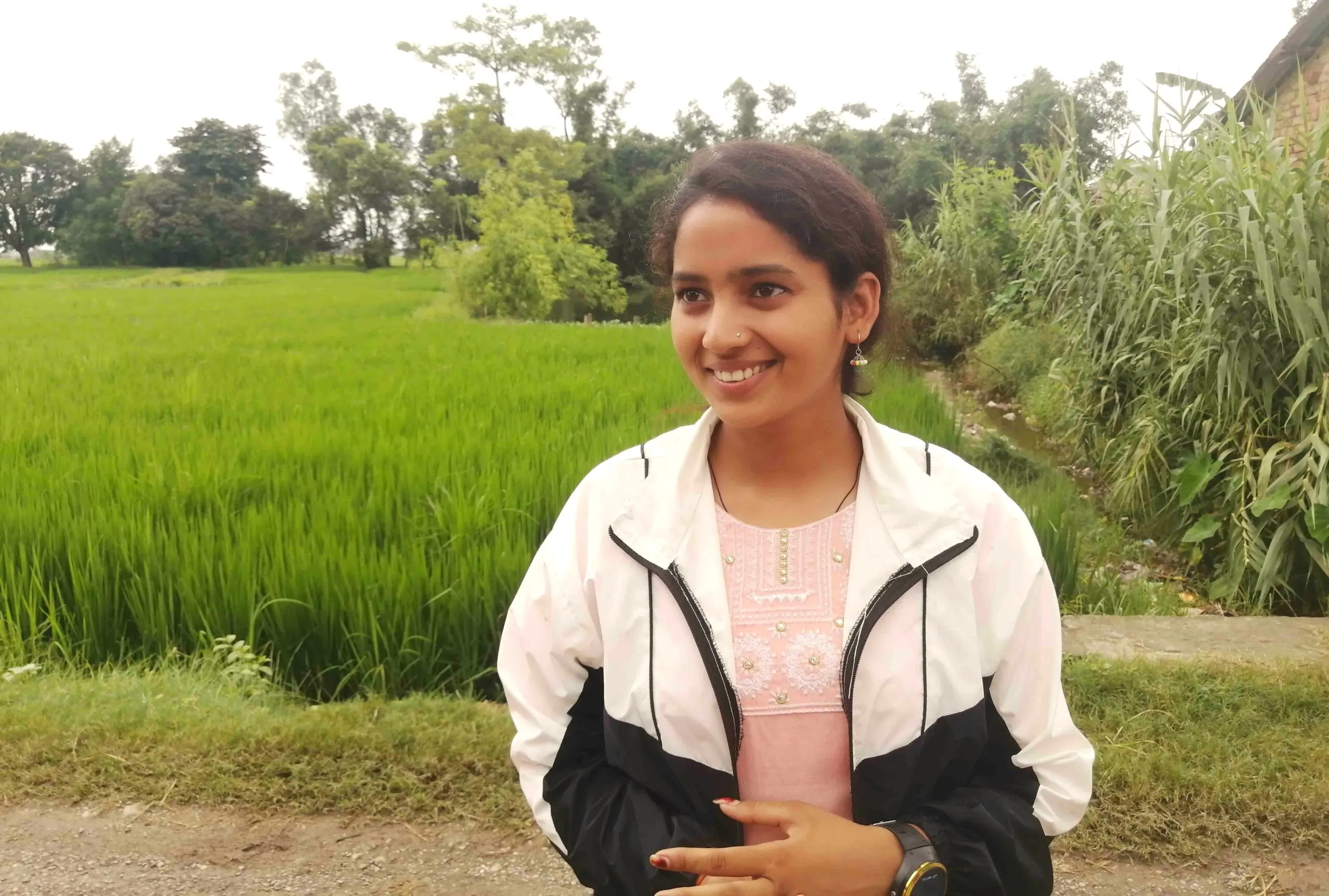 A young Nepalese girl wearing a traditional kameez tunic and a light jacket, standing in a rural setting with a paddy field in the background, looking sideways and smiling beautifully at the camera.