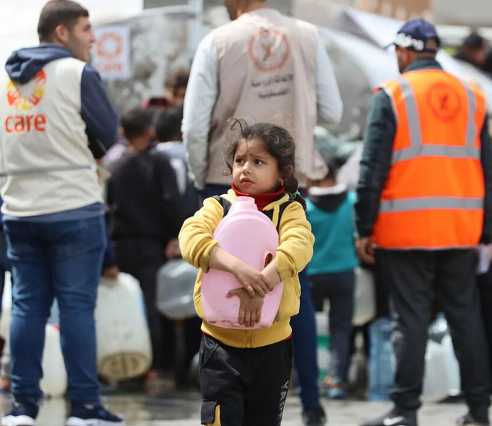 A young girl holds a pink jug close to her chest. In the background are emergency aid workers, including a man in a CARE vest.