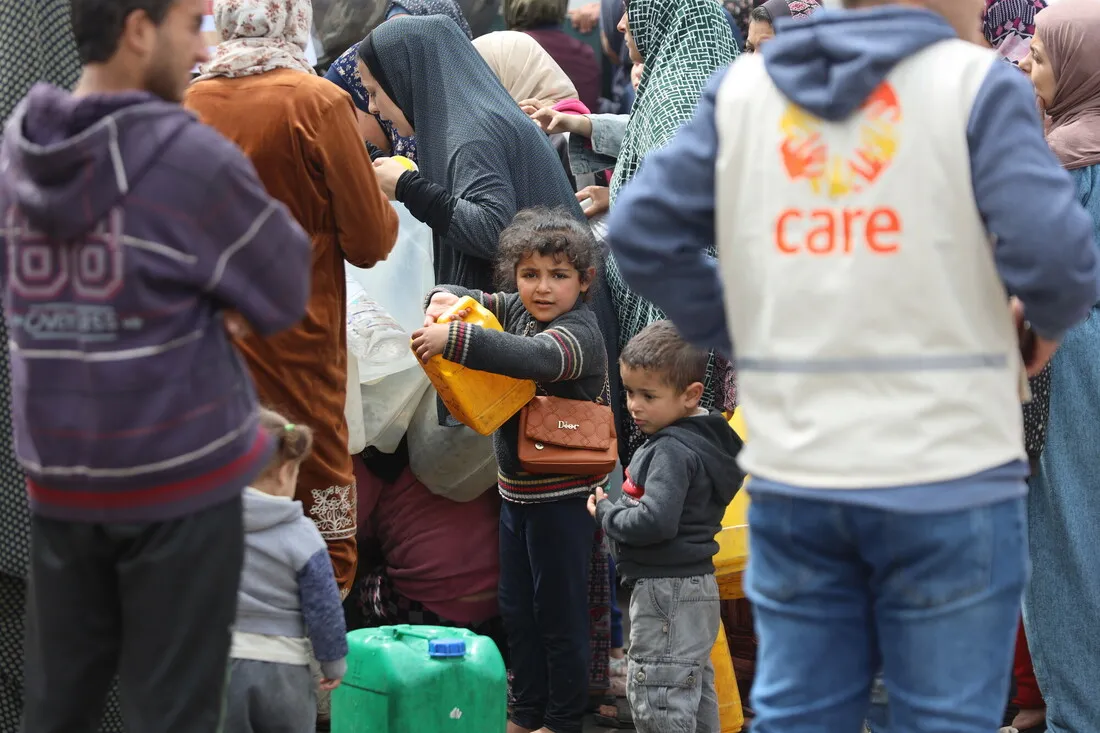 Duas crianças pequenas estão juntas, uma segurando um galão amarelo de água. Um homem vestindo um colete CARE está ao lado deles.