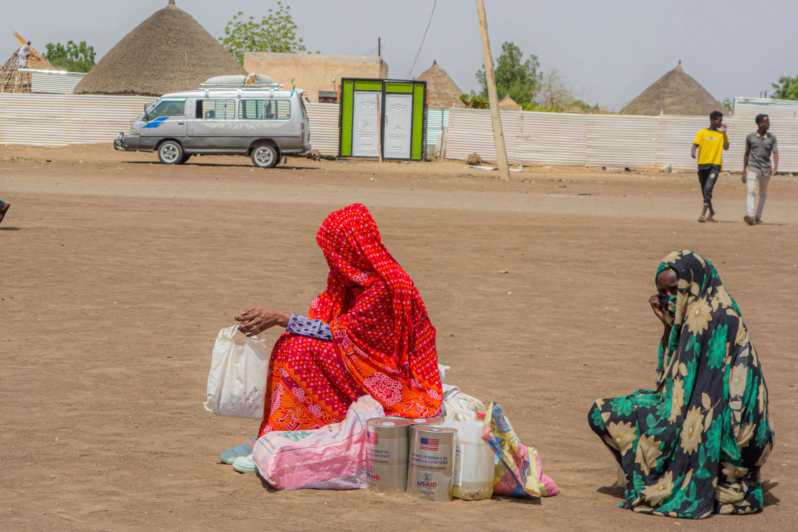 Two internally displaced Sudanese women sit on food aid sacks received from CARE, waiting for transport. One woman's face is partially visible, while the other's is fully covered. A van and two men walking are visible in the background