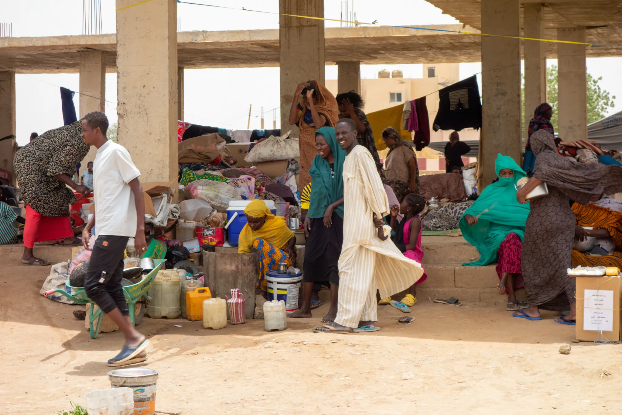 A group of internally displaced Sudanese women and men seek refuge in an unfinished building structure in Kassala, after fleeing conflict in Sinnar and Sinjah. They carry small packs and boxes containing the few basic belongings they were able to take with them as they escaped the violence.