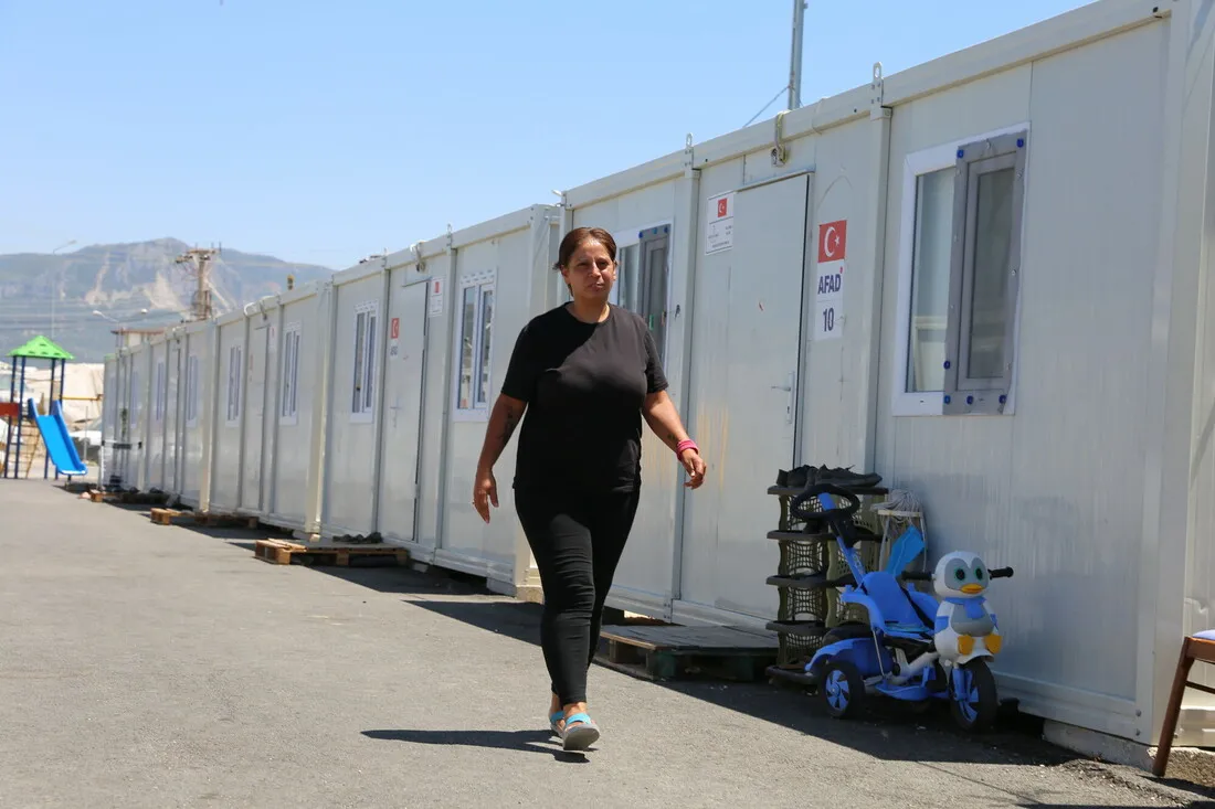 A middle-aged Turkish woman, Elcin, walks confidently towards the camera along a row of container homes. The scene symbolizes the resilience of earthquake survivors like Elcin, who are rebuilding their lives in temporary shelters.