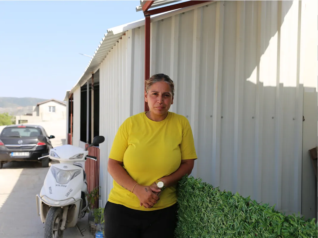 A middle-aged Turkish woman stands confidently in front of a row of container homes, where she has lived since the February 2023 earthquake. She looks directly at the camera, with a scooter parked beside her and a car in the background. Despite the bright sunny day, she stands in the shadow of the container, exuding calm determination amidst her challenging circumstances.