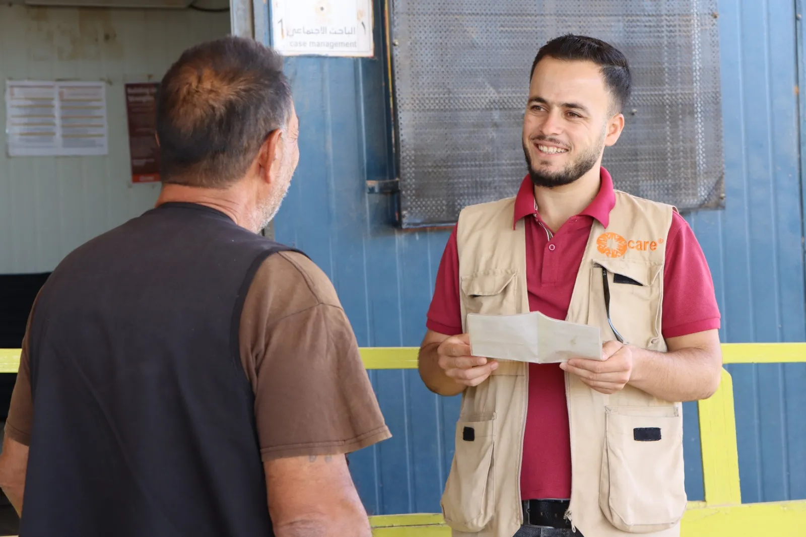 A Syrian man wearing a CARE vest, representing CARE Jordan, smiles and engages in conversation with a refugee while holding some papers.