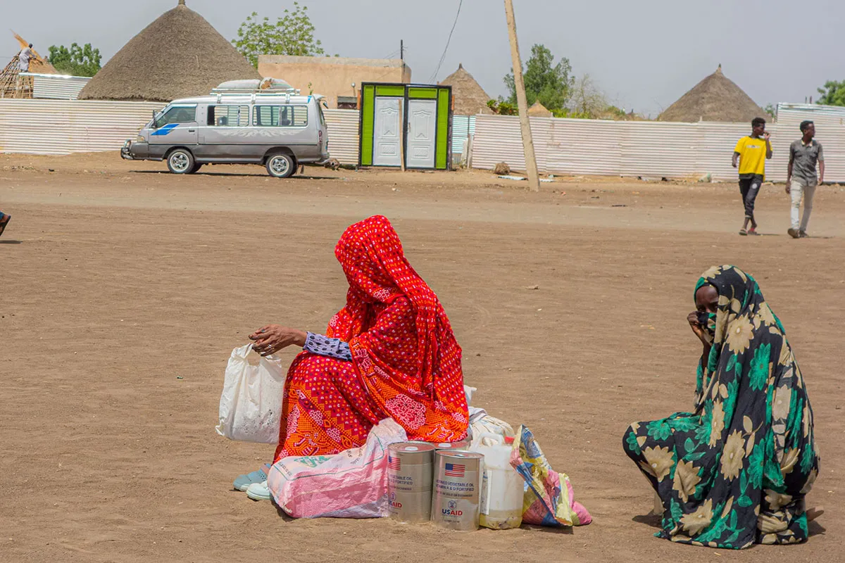 Two internally displaced Sudanese women sit on food aid sacks received from CARE, waiting for transport. One woman's face is partially visible, while the other's is fully covered. A van and two men walking are visible in the background