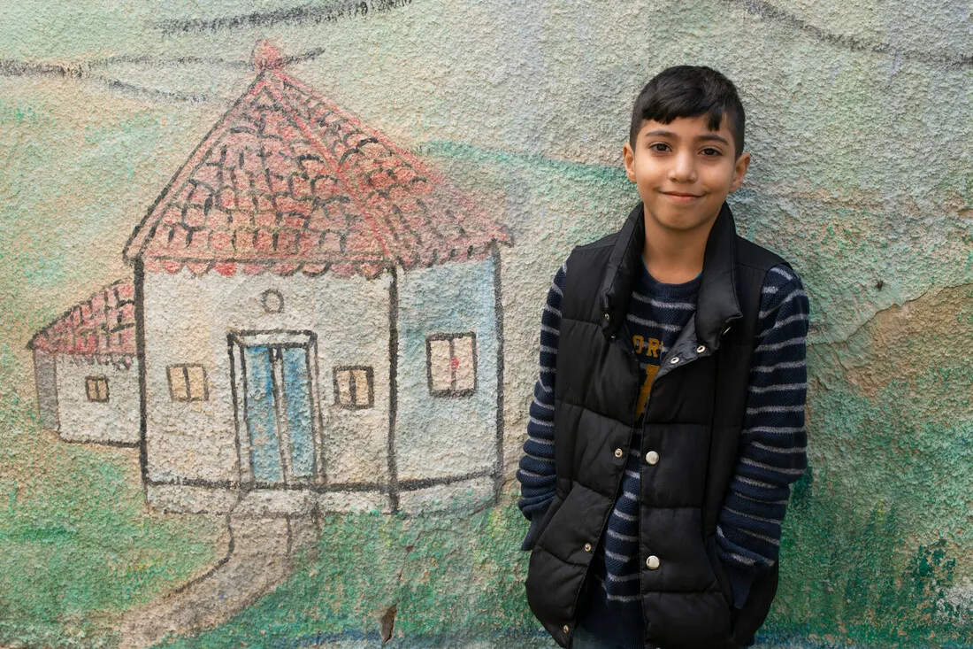 Sahwan, a 9-year-old Syrian refugee, smiles at the camera with his hands in the pockets of his puffer jacket. Behind him, a beautiful drawing of a house on the wall symbolizes the longing of Syrians for their lost homes.