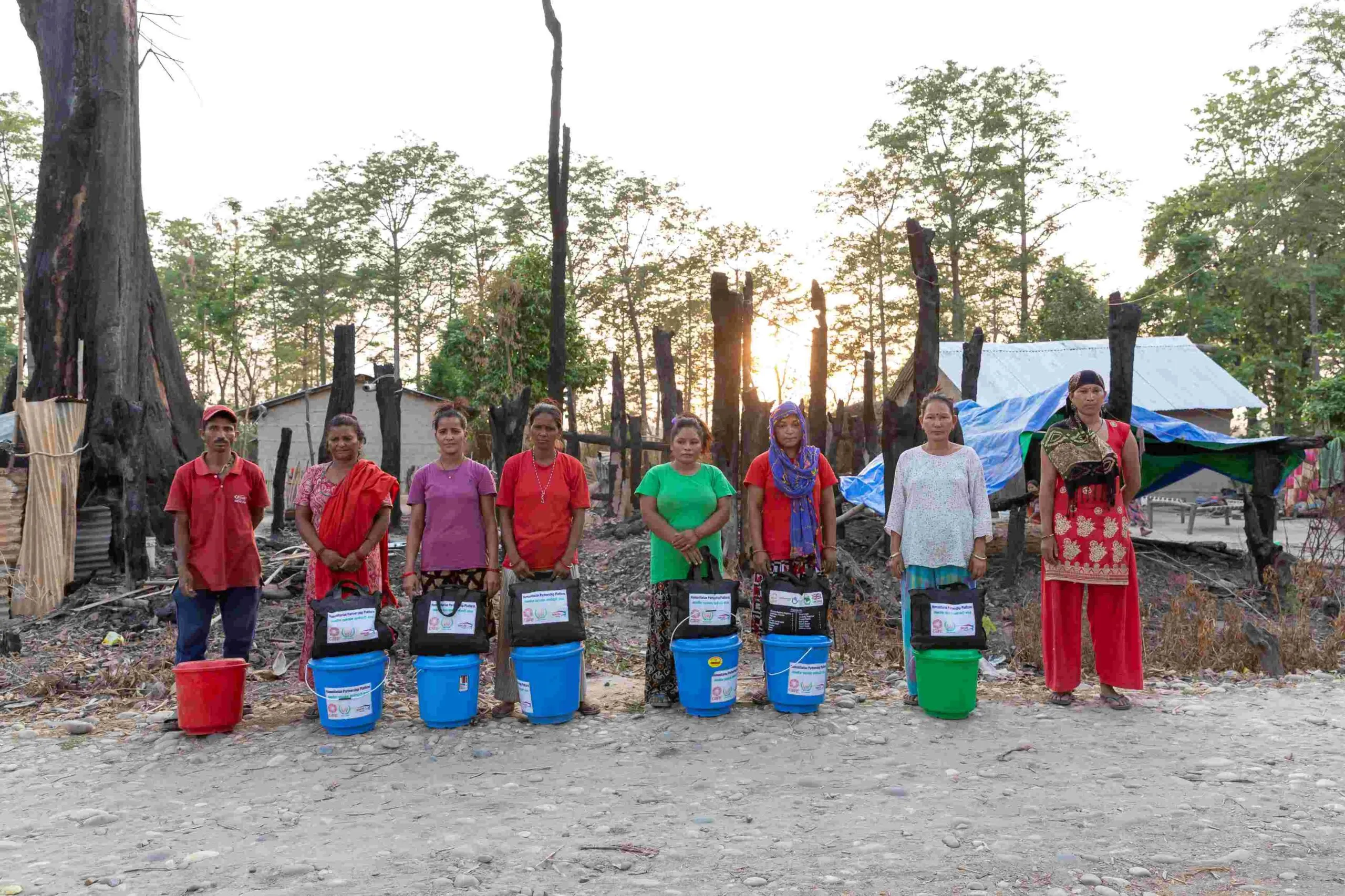 A group of eight individuals, seven women and one man, standing in a line and posing for the camera. In front of them are large plastic buckets, likely containing essential daily items, along with square-shaped packets that may hold a blanket or mosquito net.