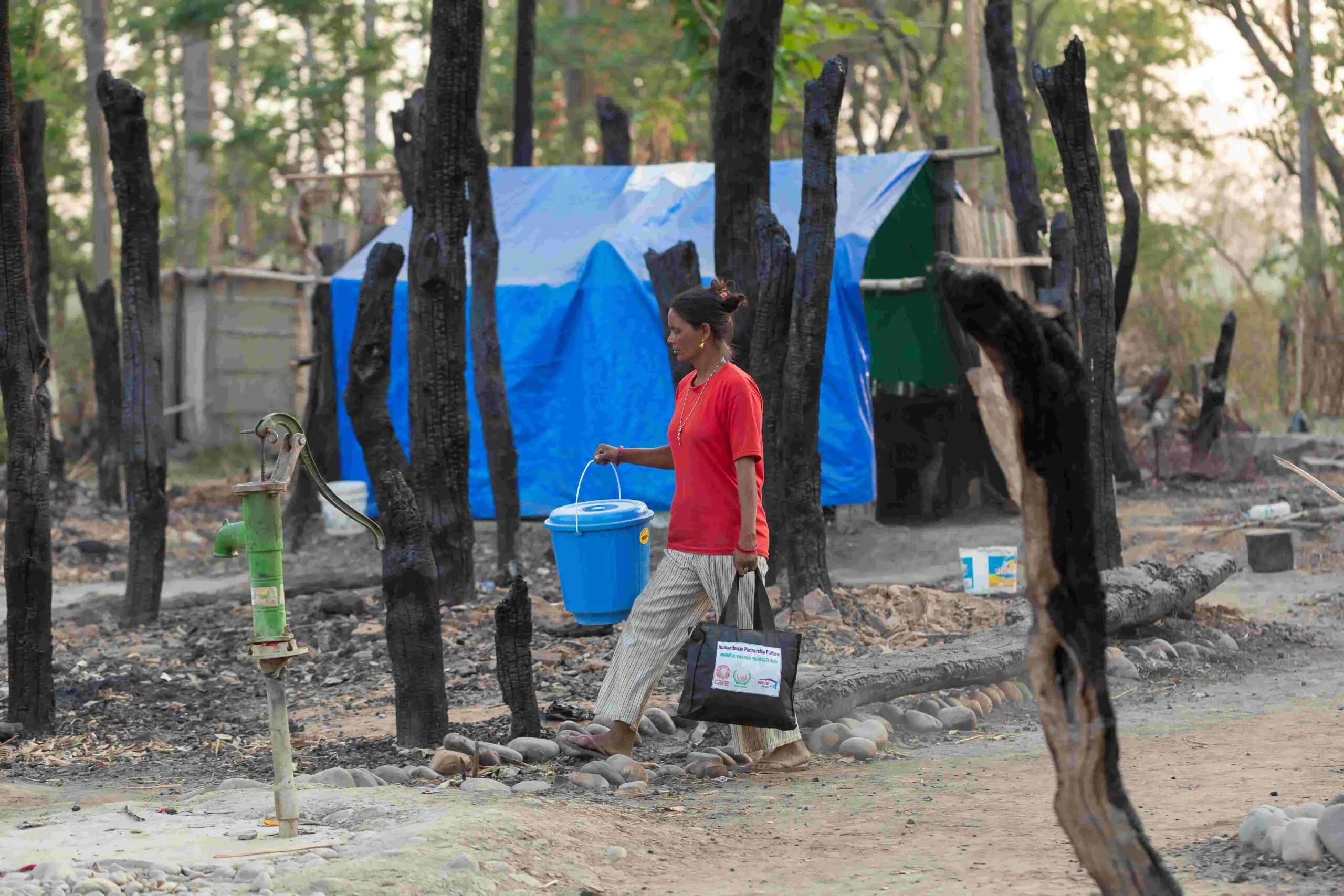 A scene of devastation shows charred black trees engulfed by fire, with makeshift shelters made of tarpaulin visible in the background. A woman walks by, carrying essential items in her hands, likely humanitarian aid or non-food supplies urgently needed after the fire incident.