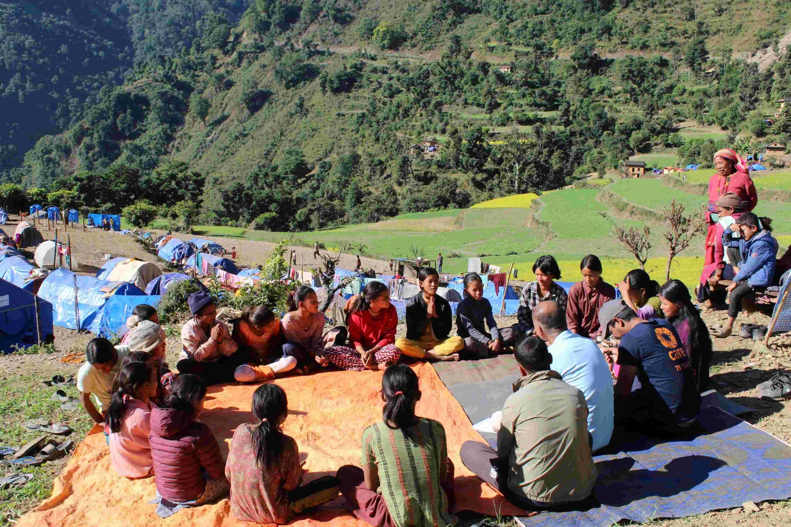 A CARE team is engaging in conversation with local community members, primarily young women, as part of a Rapid Gender Assessment following the 2023 earthquake. They are seated on a mat under the open sky in Jajarkot district, Nepal, with makeshift tarpaulin shelters visible in the background.