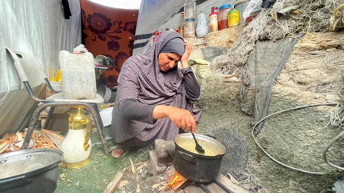 A displaced mother, sitting outside a makeshift tent, cooks a meal over an open fire. She looks concerned and worried.