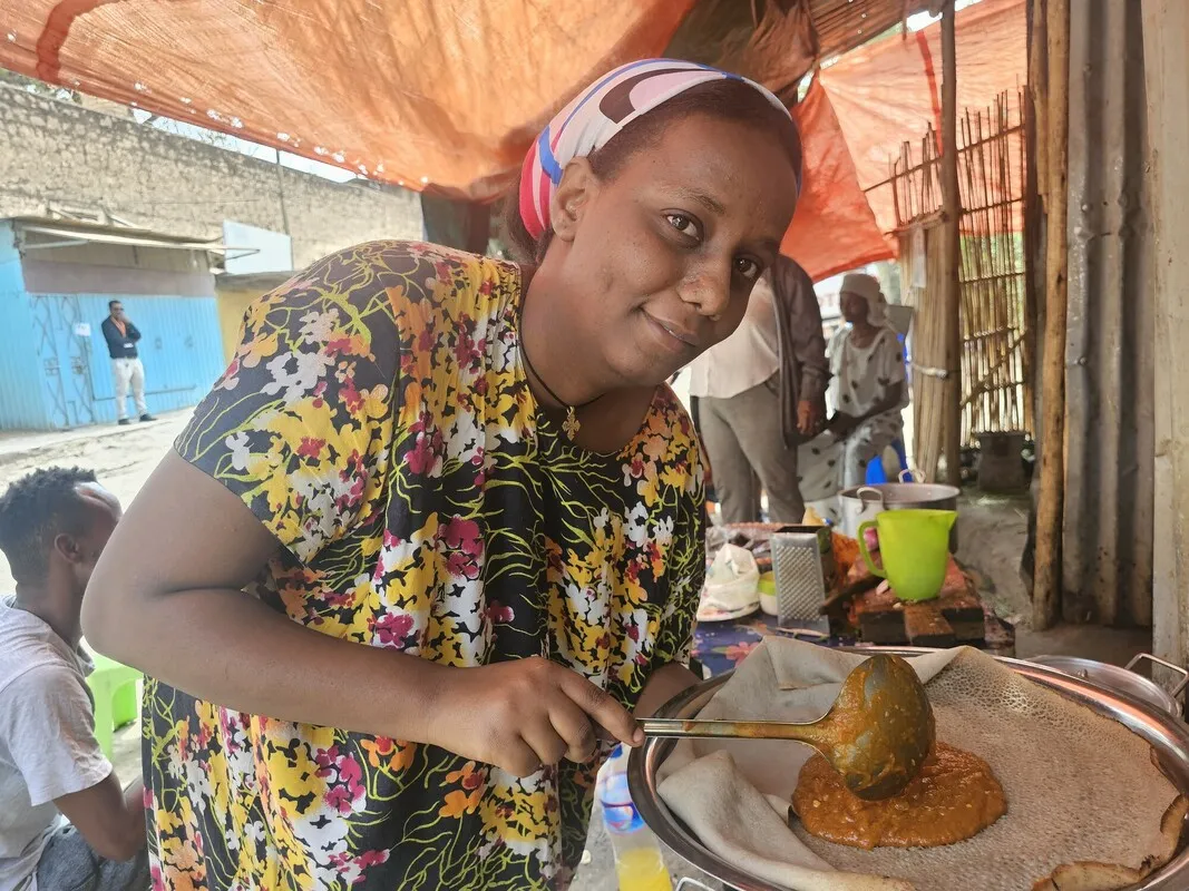 An Ethiopian woman, gazing at the camera woman, prepares a traditional meal in her food shop.