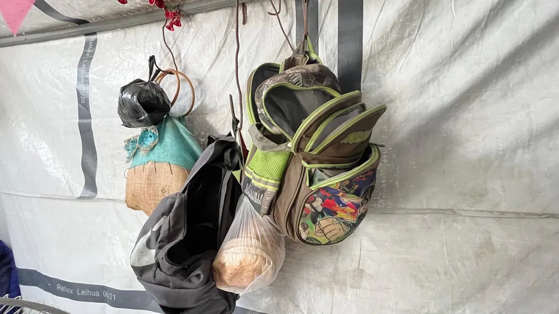 A displaced family's belongings, including two backpacks and a few plastic bags, hang on the wall of their temporary shelter, a tent.