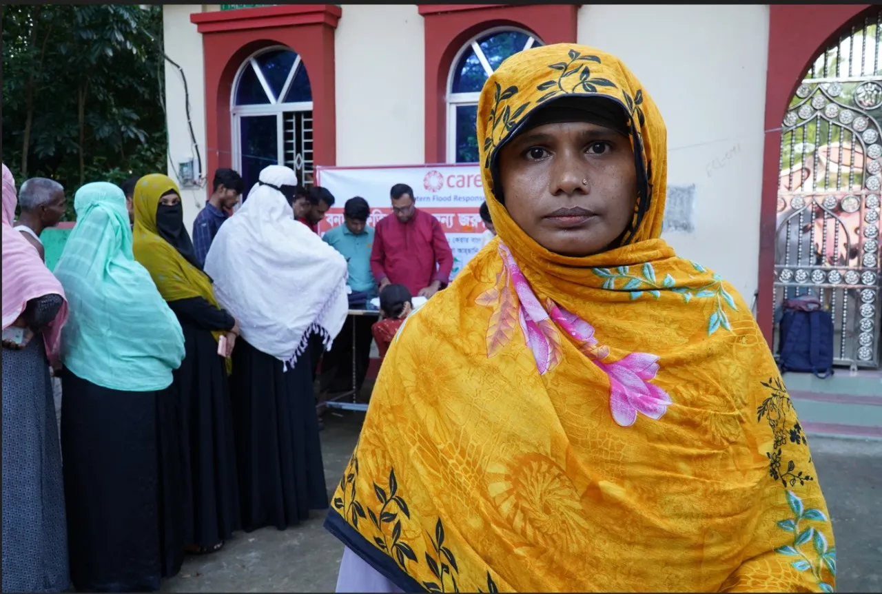 Rozina, a flood-affected woman, gazes at the camera and stands near a queue of women receiving relief assistance from CARE. A CARE banner is visible in the background.