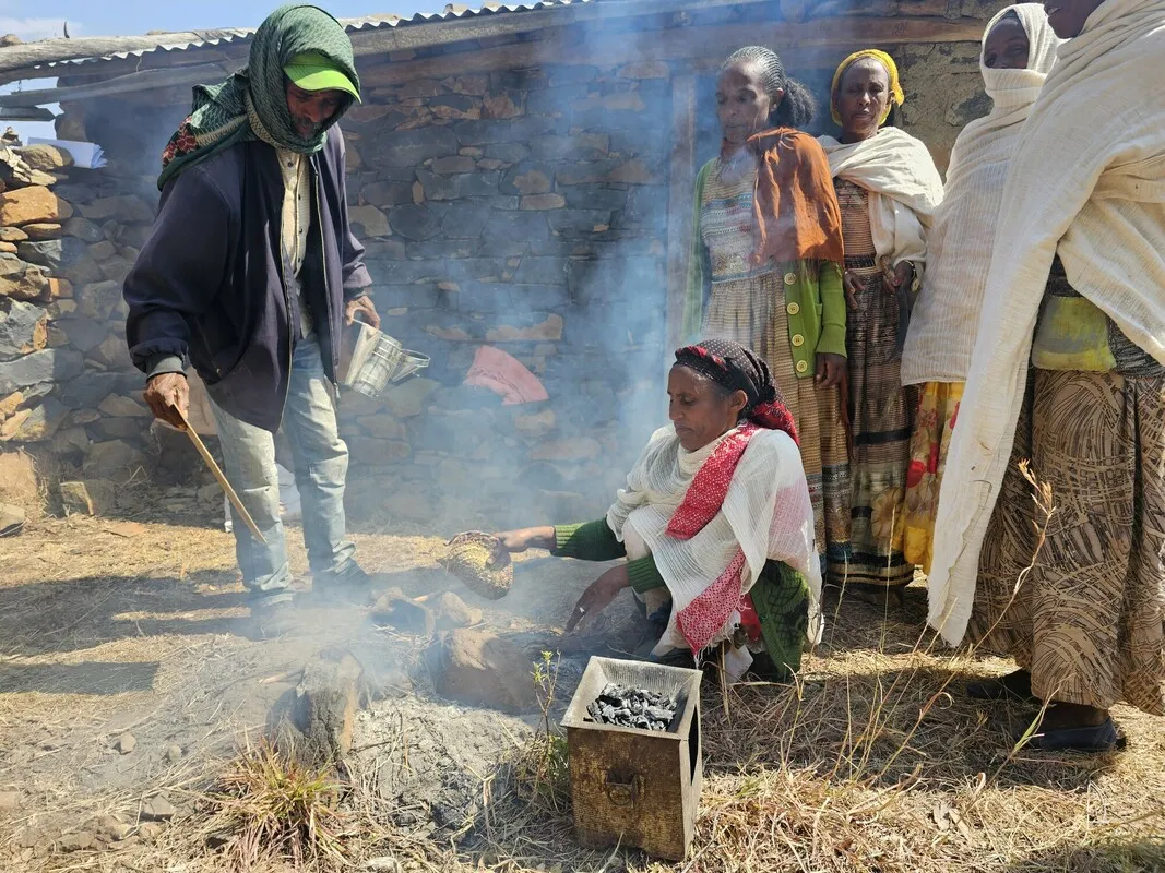 A man standing and a woman sitting are creating smoke to calm the bees. Four other women, possibly members of a beekeeping cooperative, watch intently.