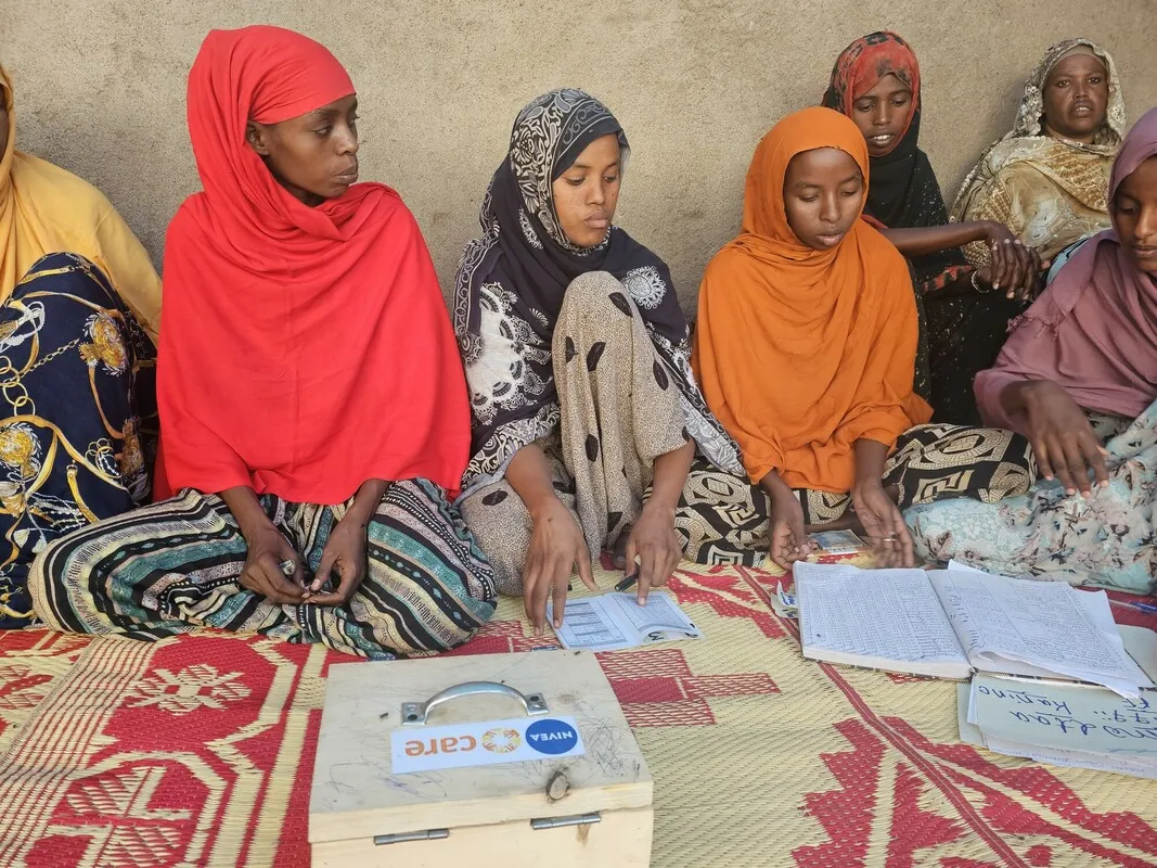 A savings group meeting with women focused on their notebooks and accounts. A wooden savings box with a CARE sticker is prominently displayed.