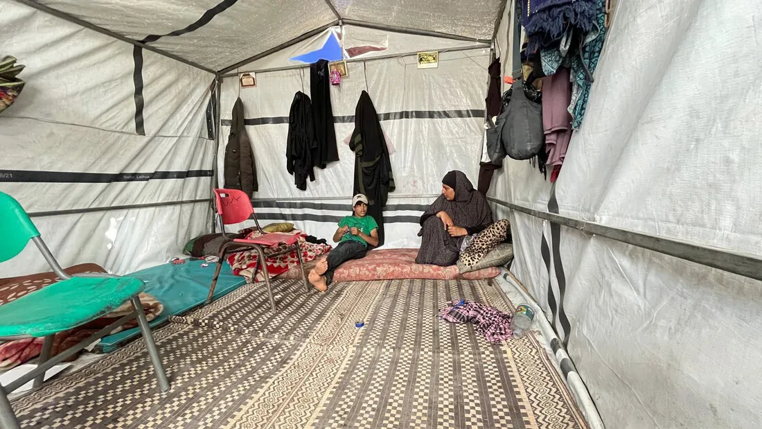 A displaced mother and her son having a chat inside their makeshift tent home where a few belongings, including clothes and bags, hang on the tarpaulin walls, and mattresses and pillows on the floor.