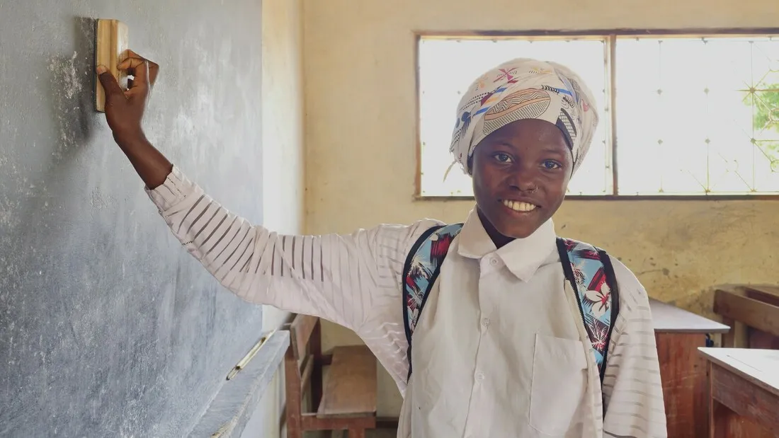 Farsana, a girl from northern Mozambique, smiling at the camera and cleaning the blackboard in her classroom.