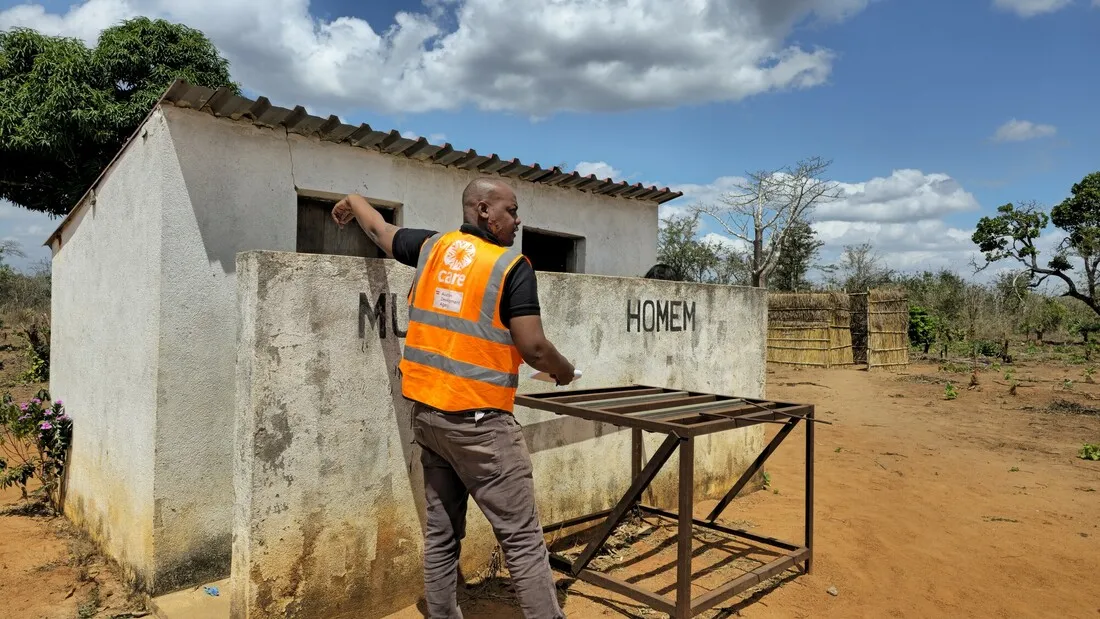 A person wearing a CARE vest is pointing towards a school latrine that features signage indicating separate male and female toilets.