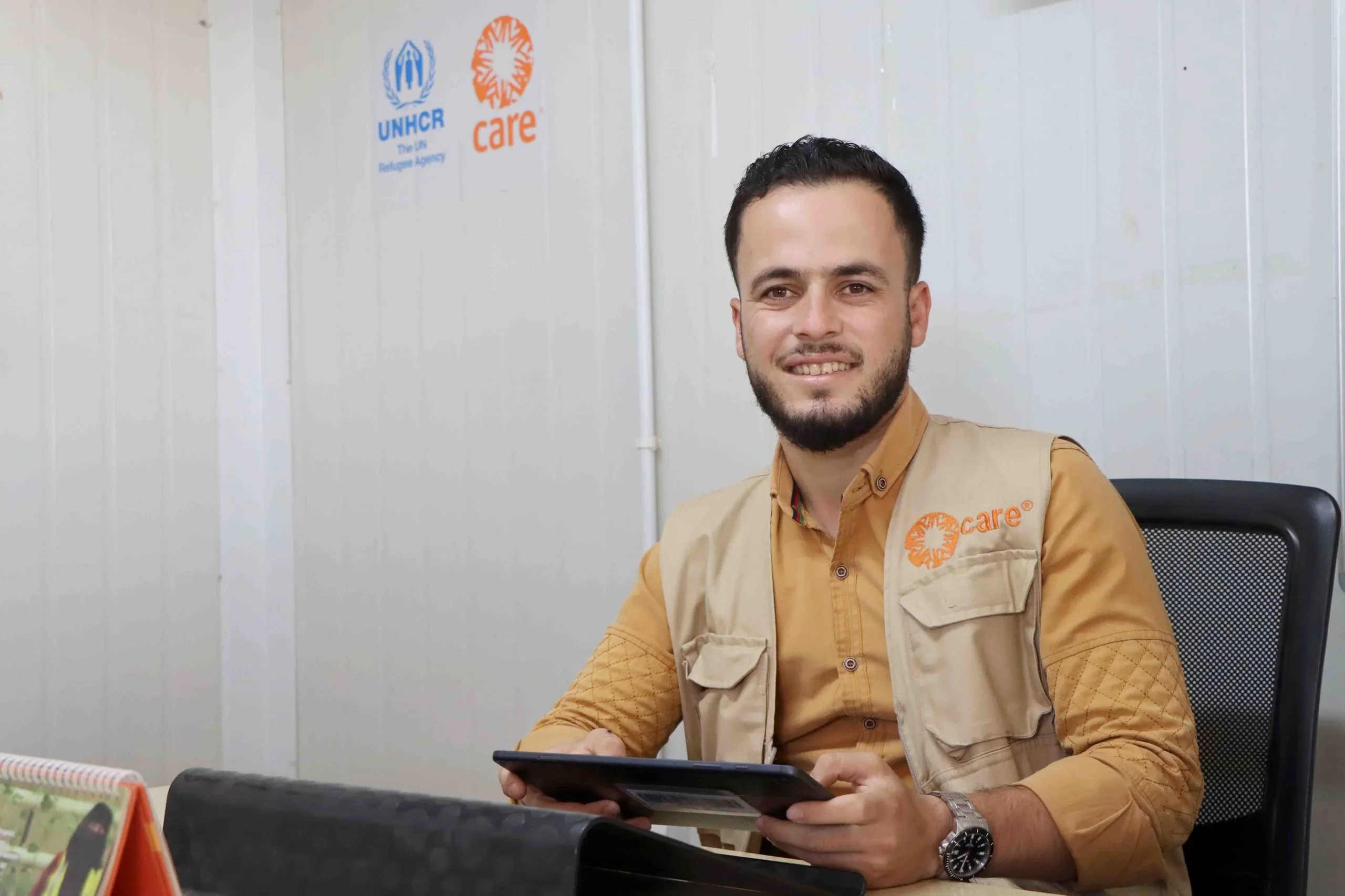 A Syrian man wearing a CARE vest gazes at the camera in a CARE Jordan office, with the CARE logo displayed in the background.