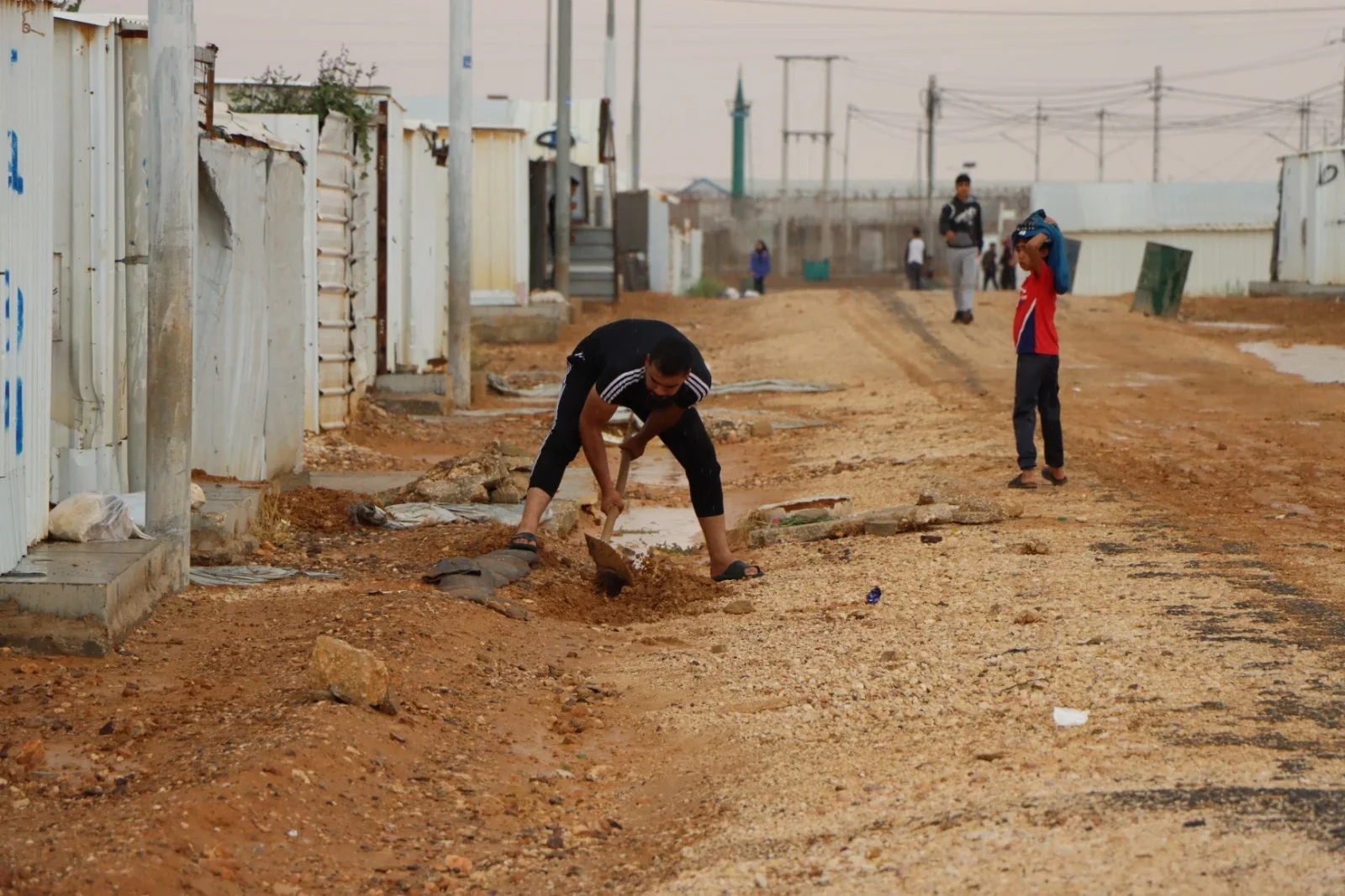 Long rows of white container-like houses fill a refugee camp, where a man, likely a refugee, digs a sewage channel with a spade. A young boy pauses nearby to observe him, while a group of men and women can be seen in the background, moving in different directions.