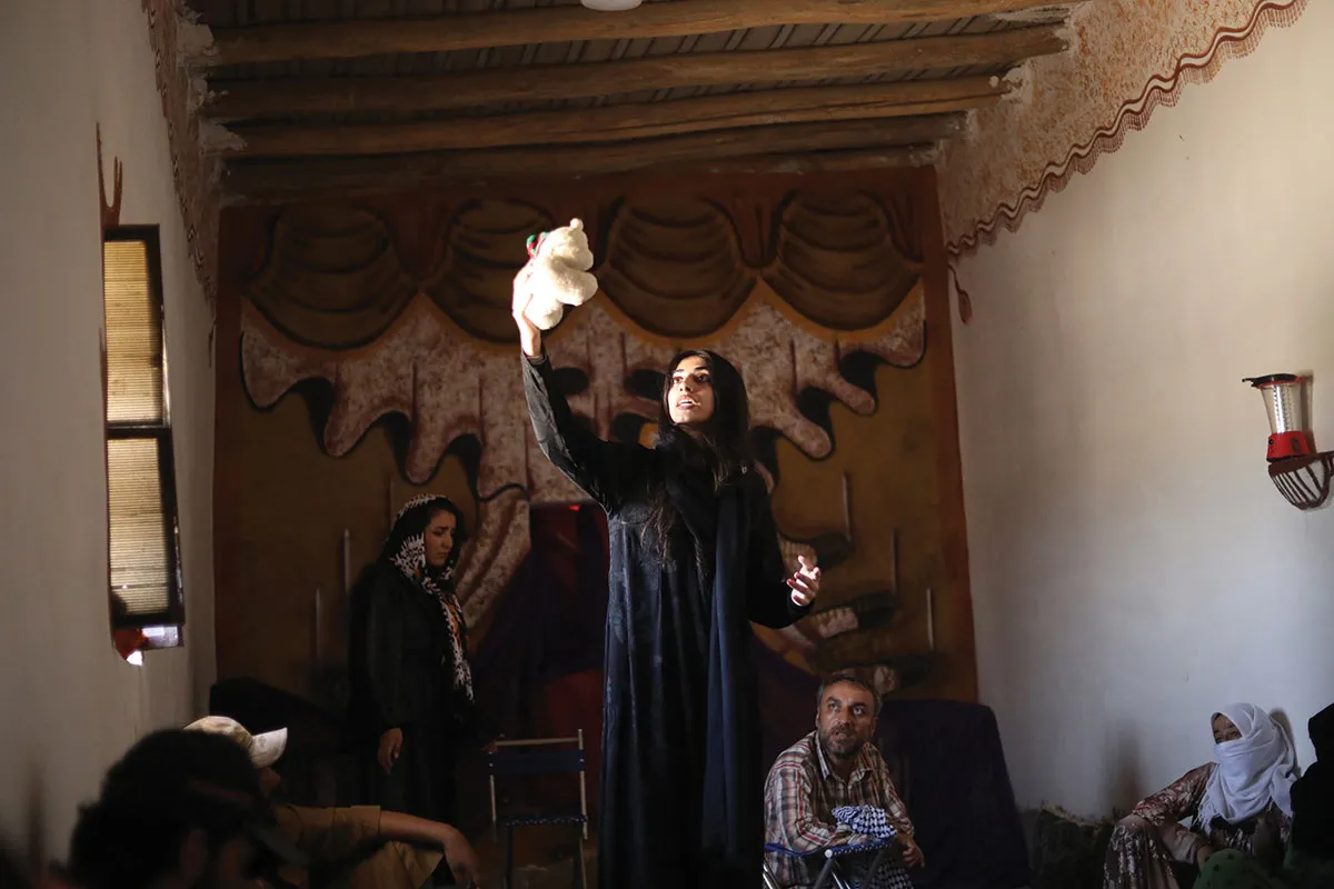 Light shines on a woman in a dark room as she acts on a stage and holds a teddy bear up in the air.