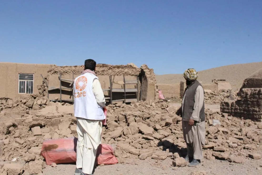 Two Afghan volunteers, one wearing a CARE vest, survey the damage to a mud home destroyed by an earthquake.